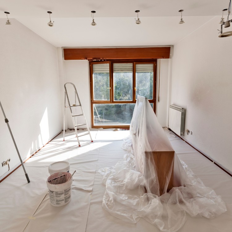 Interior room undergoing painting, featuring a covered wooden table, a ladder, paint cans, and bright natural light from large windows, highlighting Forge Construction & Design's remodeling services.