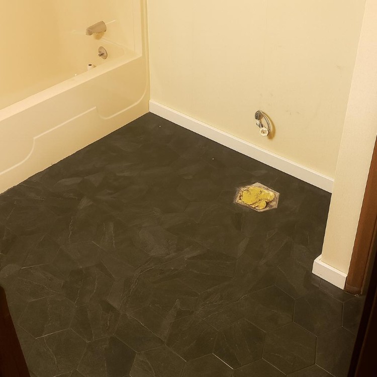 Bathroom flooring featuring hexagonal black tiles, with a bathtub in the background and a visible drain area, showcasing quality materials for home renovation in Northern Indiana.